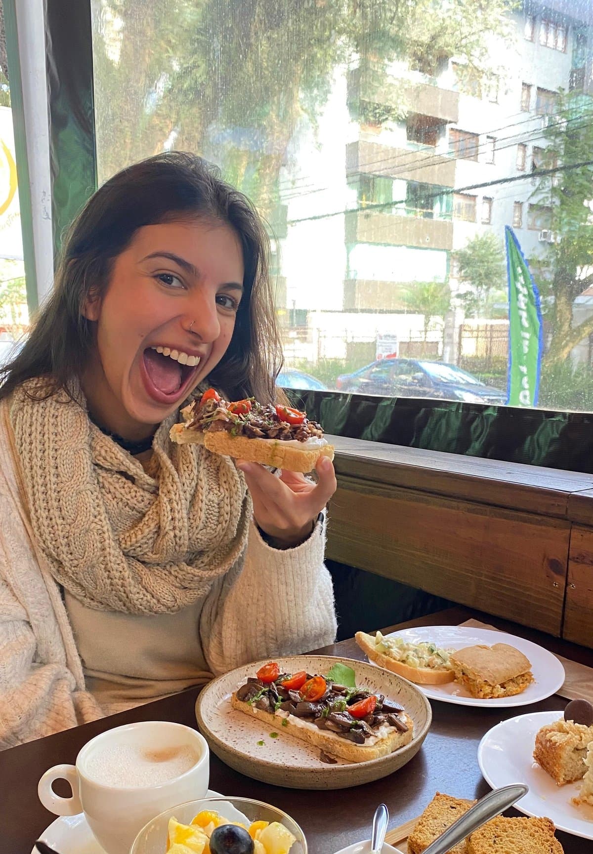 Ana Carolina Calderaro rindo enquanto come uma torrada com legumes em um café